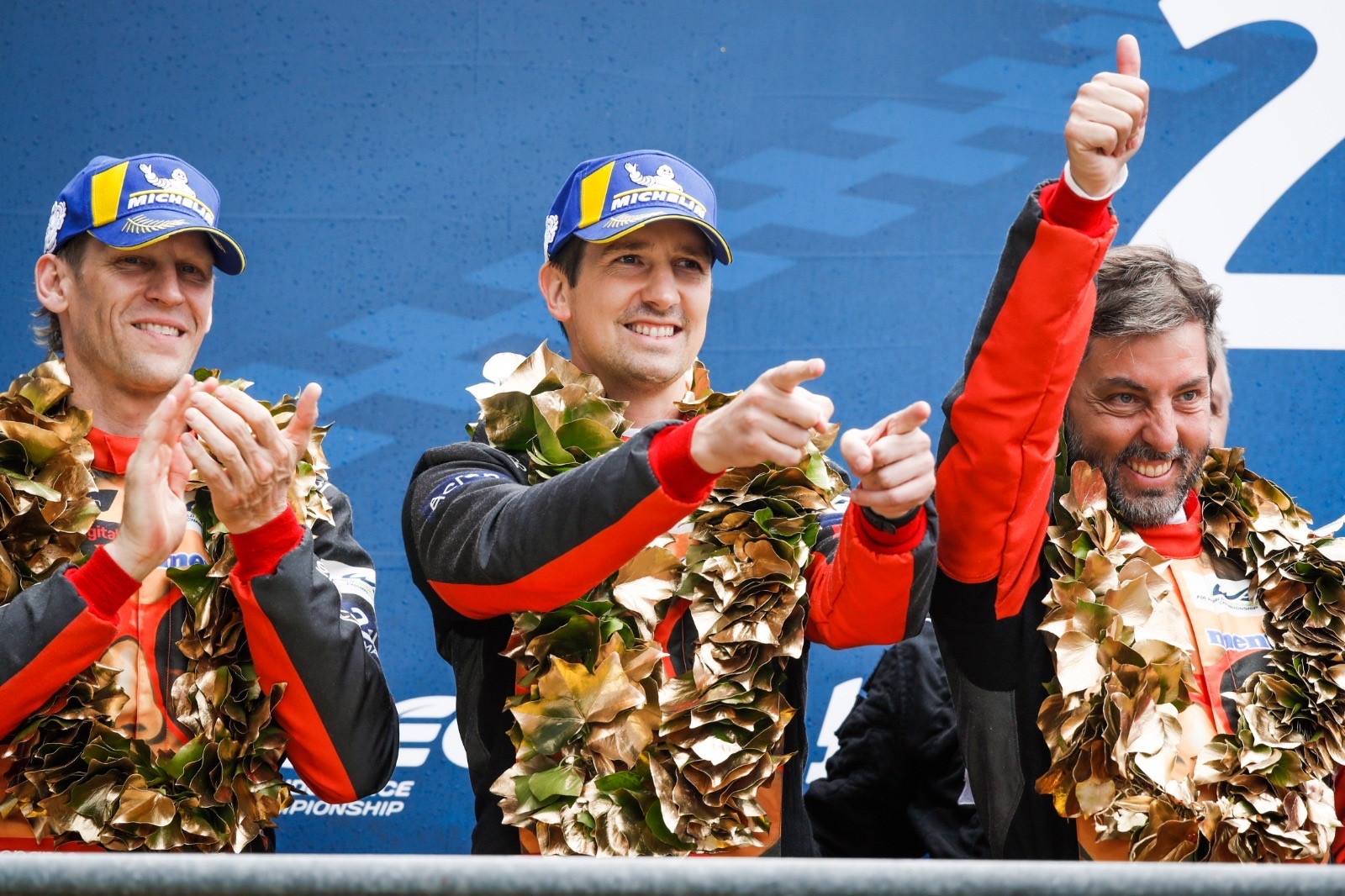 Three racers in suits and caps celebrate on a podium with wreaths, thumbs-up and pointing gestures, set against a championship backdrop.